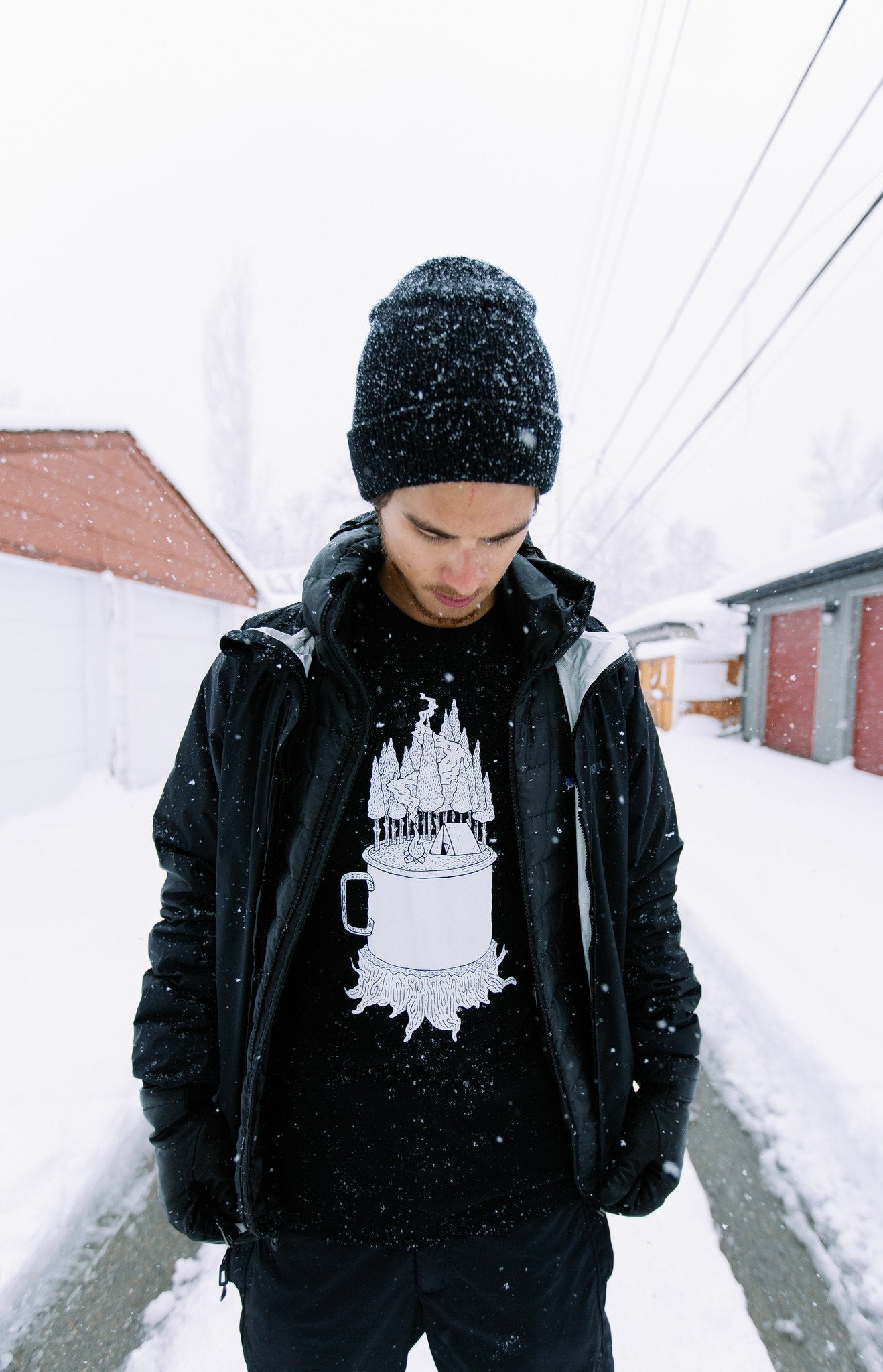 Man in the snow wearing black t-shirt, with graphic of a mug on a stump, with a camp site in the woods growing out of it.