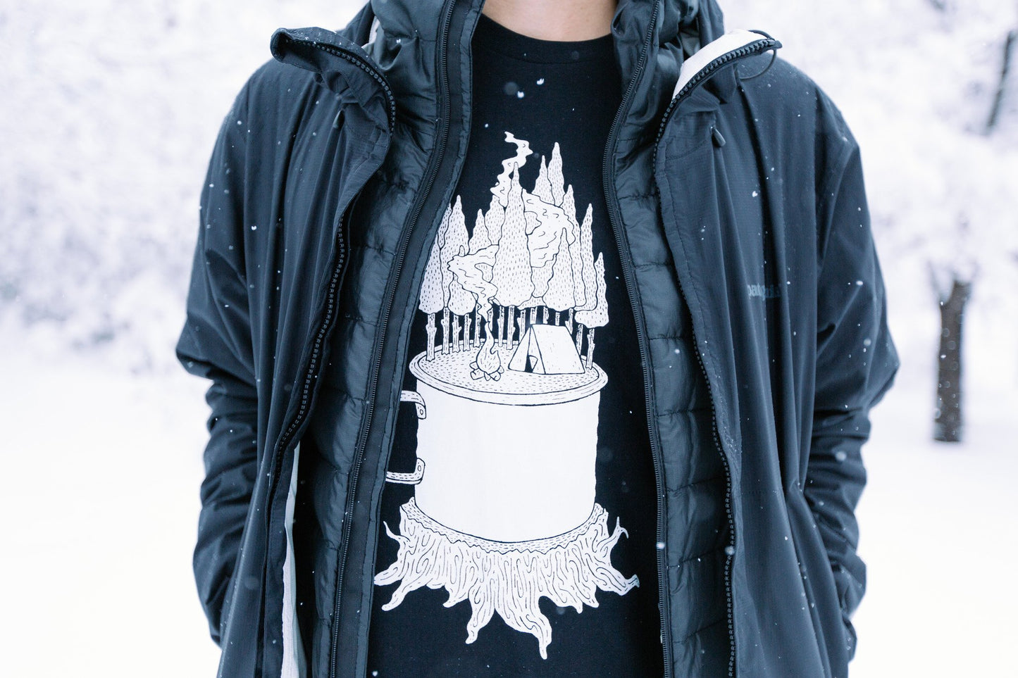 Close up of man in snow wearing black t-shirt, with graphic of a mug on a stump, with a camp site in woods growing out of it.