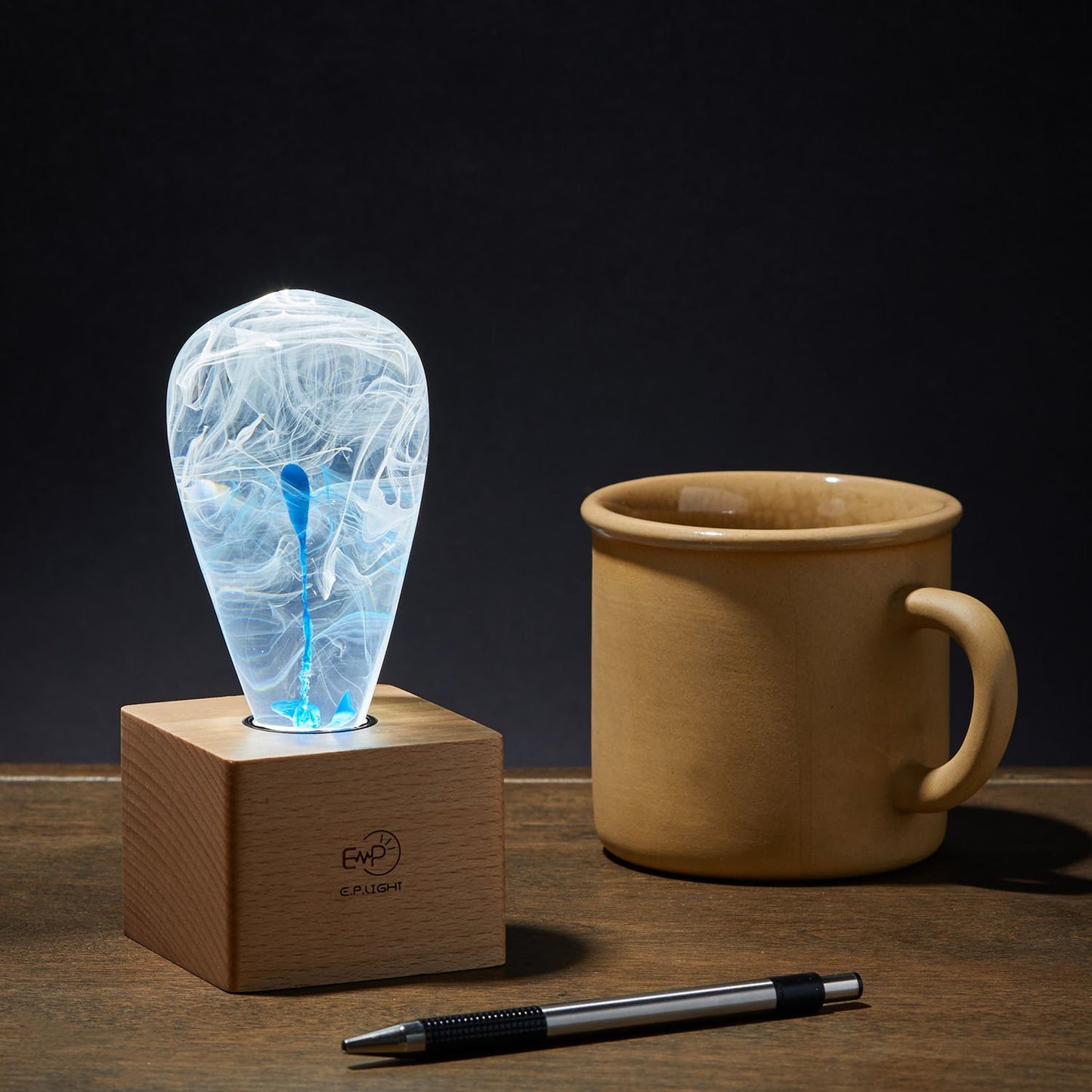 Decorative light bulb with blue and white ink drop swirl inside, screwed into a modern square wood base. Next to coffee mug.