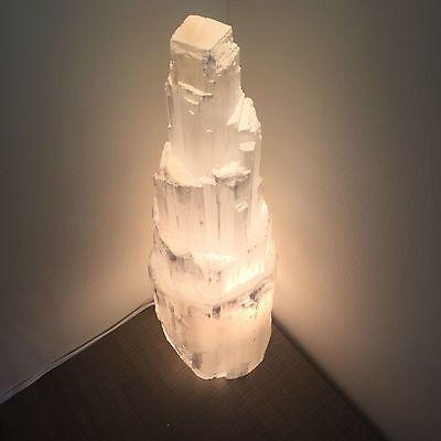 Top view of selenite crystal tower illuminated from light within, glowing in corner of dim room atop wooden table.