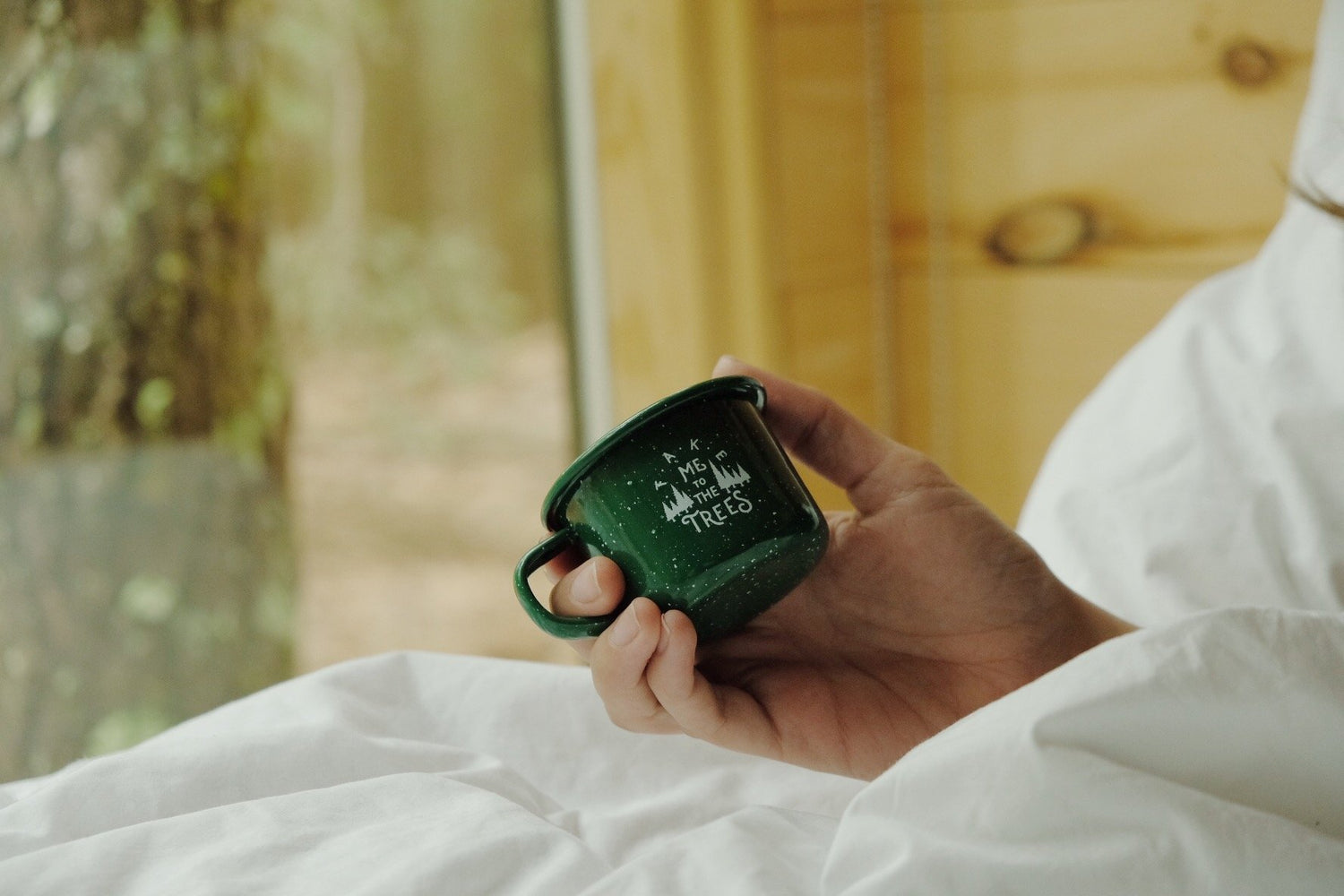 Hand holding mini green enamel mug, printed in white with graphic that says “Take me to the trees” and evergreen drawings.