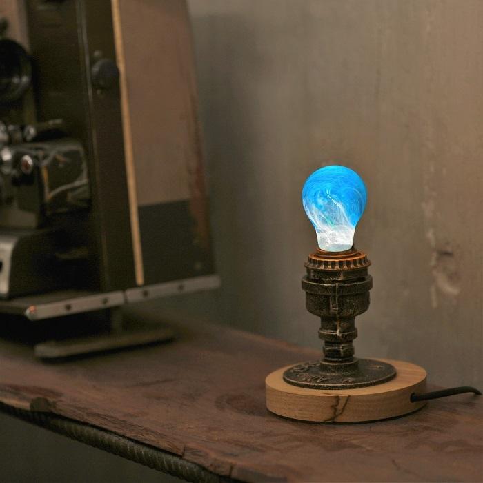 Illuminated clear lightbulb filled with blue and white swirls, in worn copper and wood base atop weathered wood shelf.