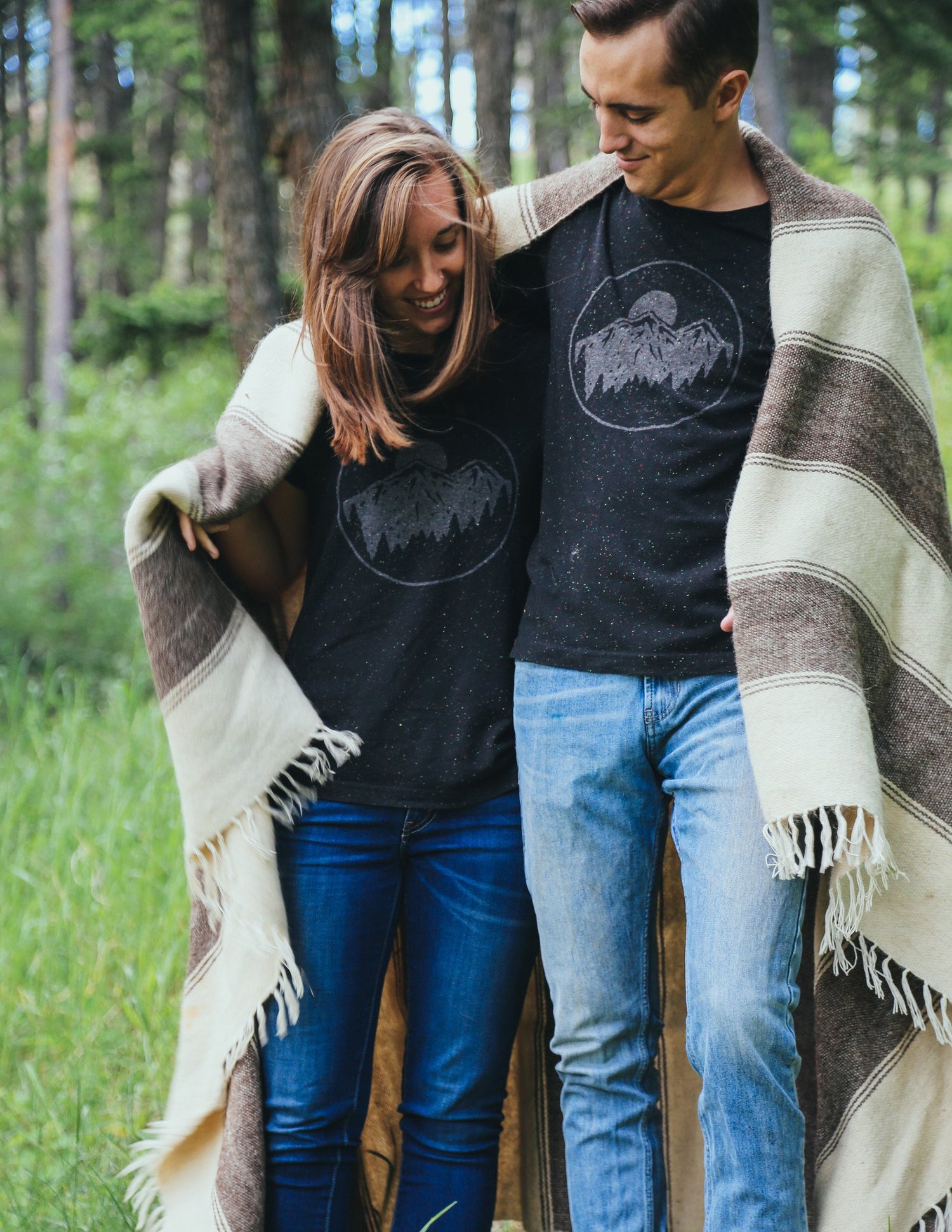 Man and woman with blanket over their shoulders, wearing matching black speckled tee shirts with moon over mountains graphic.