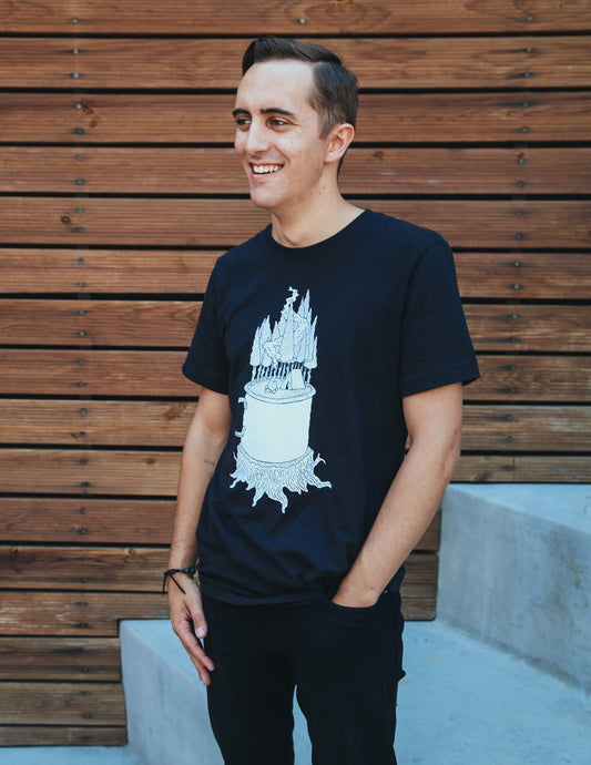 Man in a black unisex tee, with a white graphic, of a mug on top of a stump, with a camp site in the woods growing out of it.