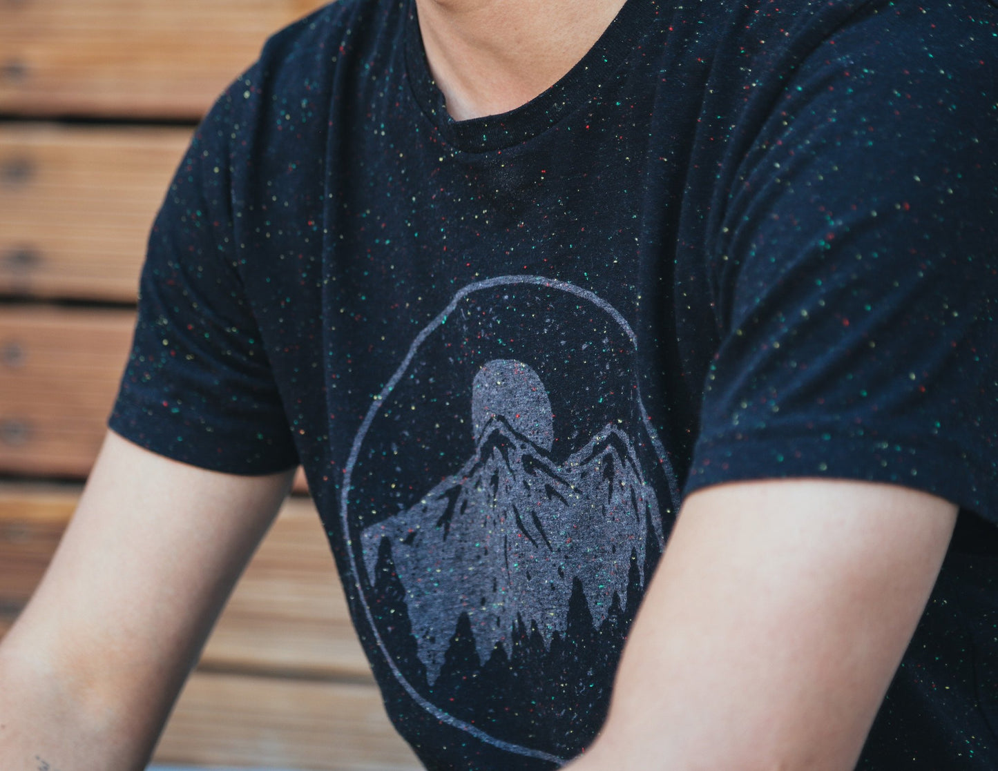 Close up from shoulder down of man in black with white speckle tee shirt, featuring moonrise over mountain range graphic.