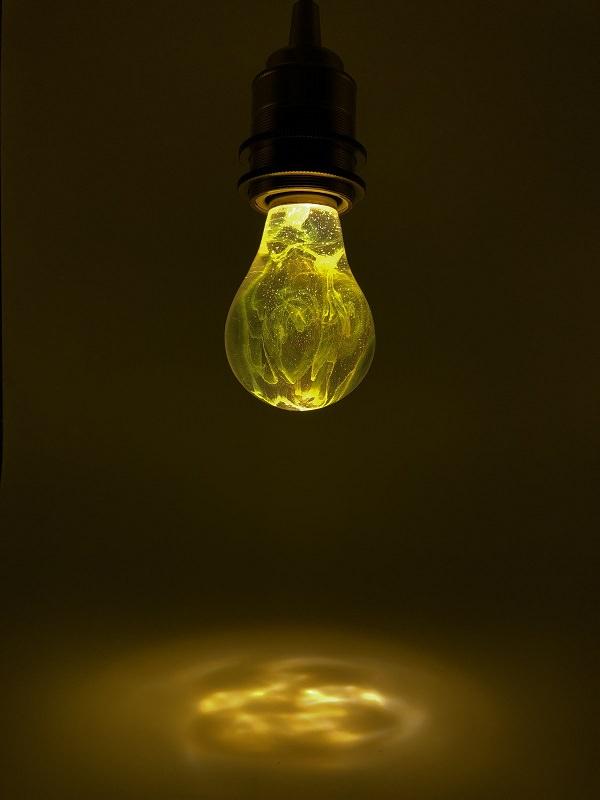 Clear lightbulb filled with yellow nebula clouds in hanging black fixture, casting warm yellow glow on white surface below.