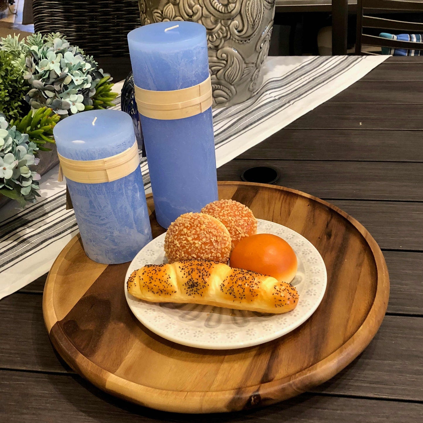 Circular acacia wood lazy susan atop dark wooden table, displaying sky blue candles and baked bread.