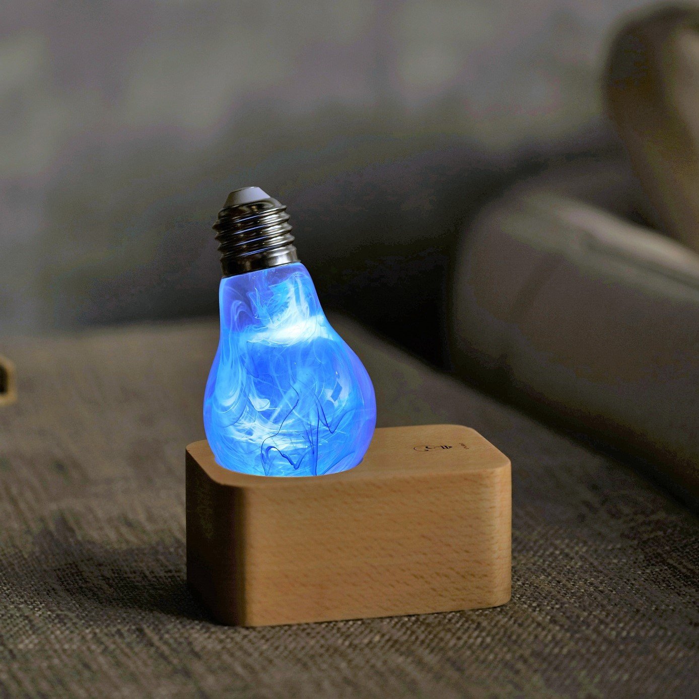 Glowing clear lightbulb filled with blue swirls, upside down in wood base atop gray fabric.