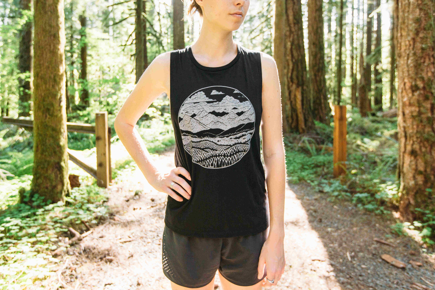 Woman in sunny woods wearing black tank top with white printed design of mountains and forest within a circle.