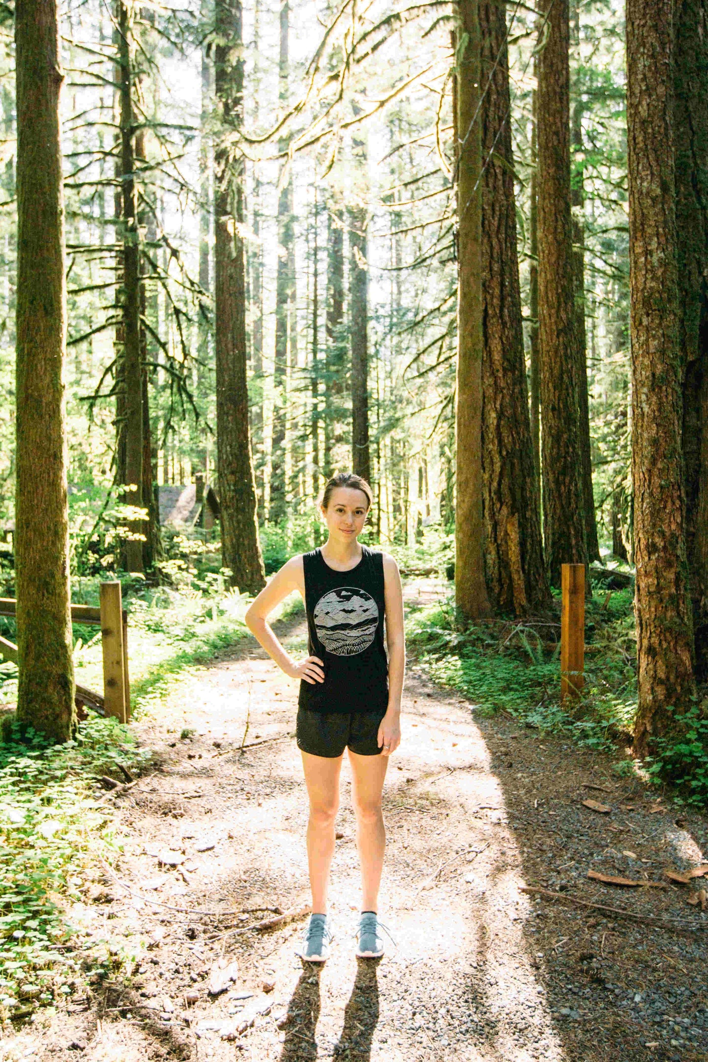 Smiling woman in sunny forest wearing gray shorts and a black tank top with white graphic of mountains and woods.