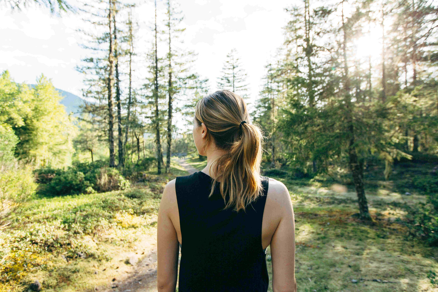 Back view of woman’s shoulders wearing black tank top in sunny clearing in woods.