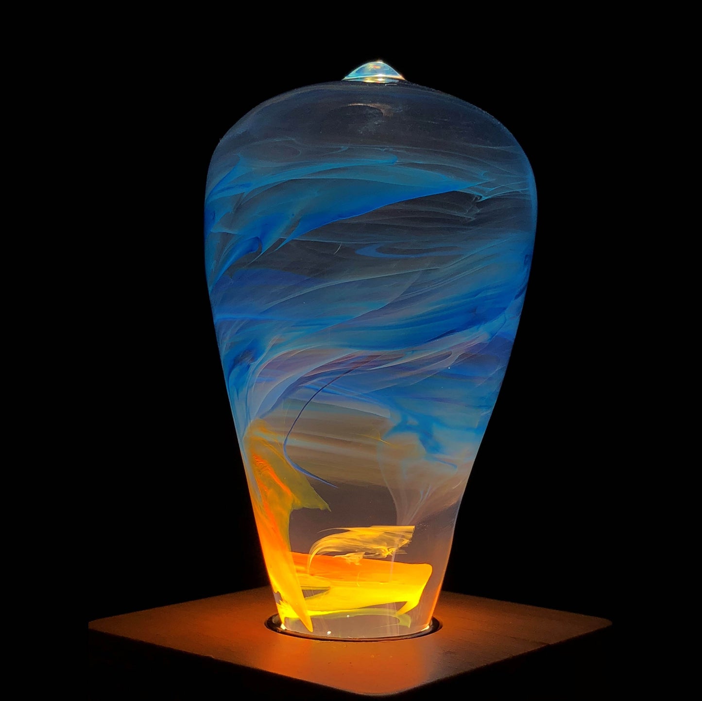 Close up view of illuminated clear lightbulb with yellow and blue swirls, set in wooden base in dark room.