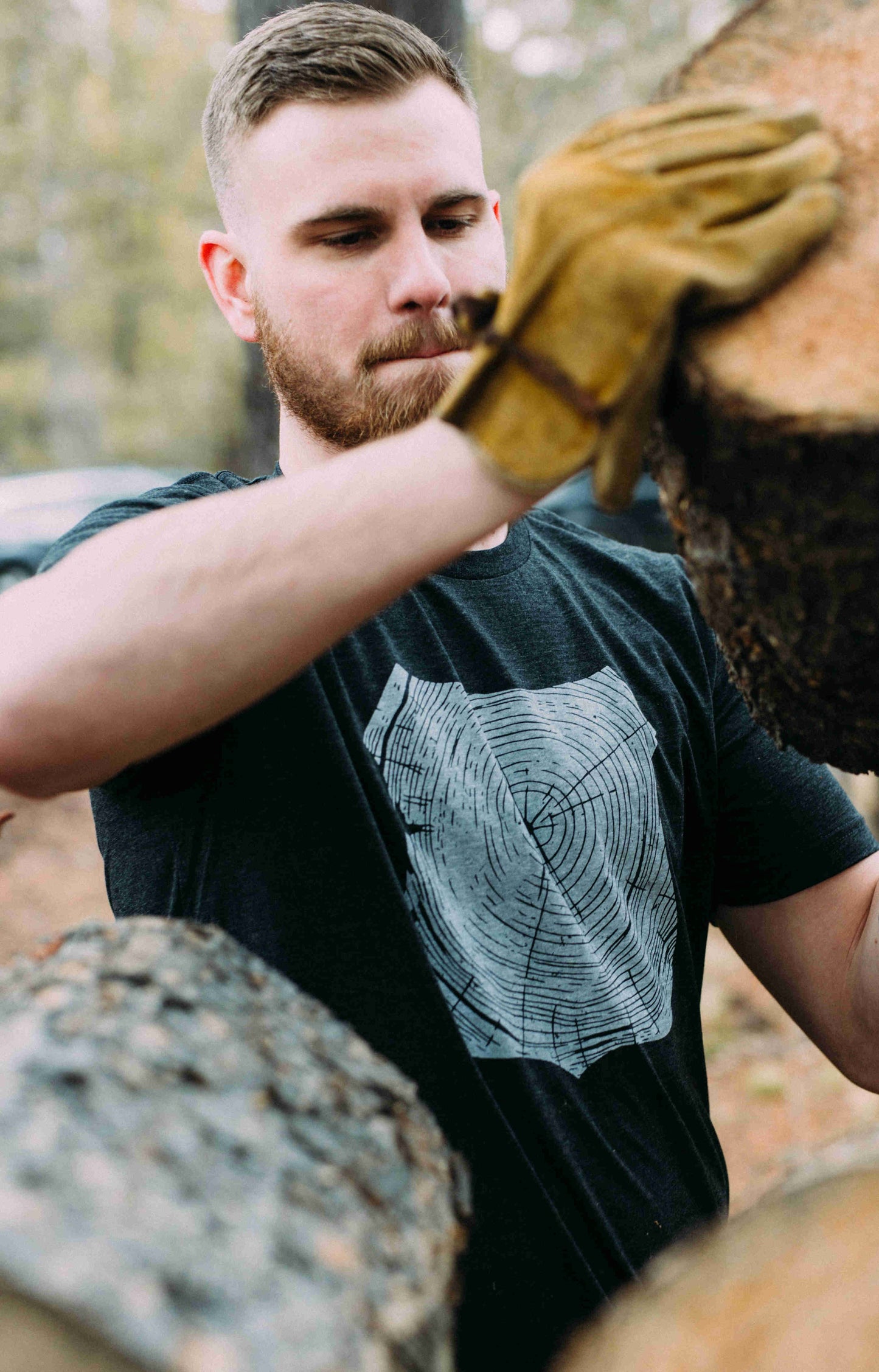 Man stacking cut logs, wearing gray tee shirt printed with white forest ranger badge outline, filled with tree rings.