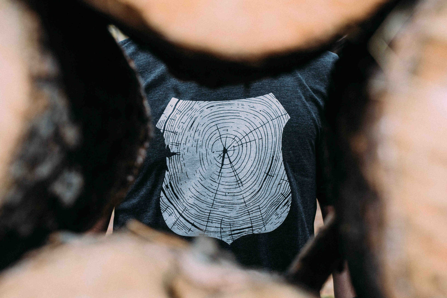 Man seen through stack of logs, wearing gray tee shirt printed with white forest ranger badge outline, filled with tree rings.