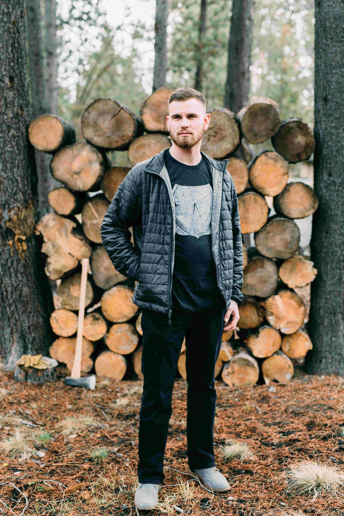 Man standing in front of cut logs, in gray tee shirt printed with white forest ranger badge outline, filled with tree rings.