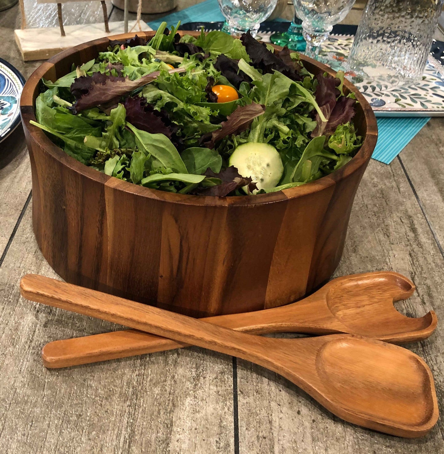 12 inch Acacia wood straight sided salad bowl filled with salad on a table next to two matching serving utensils.