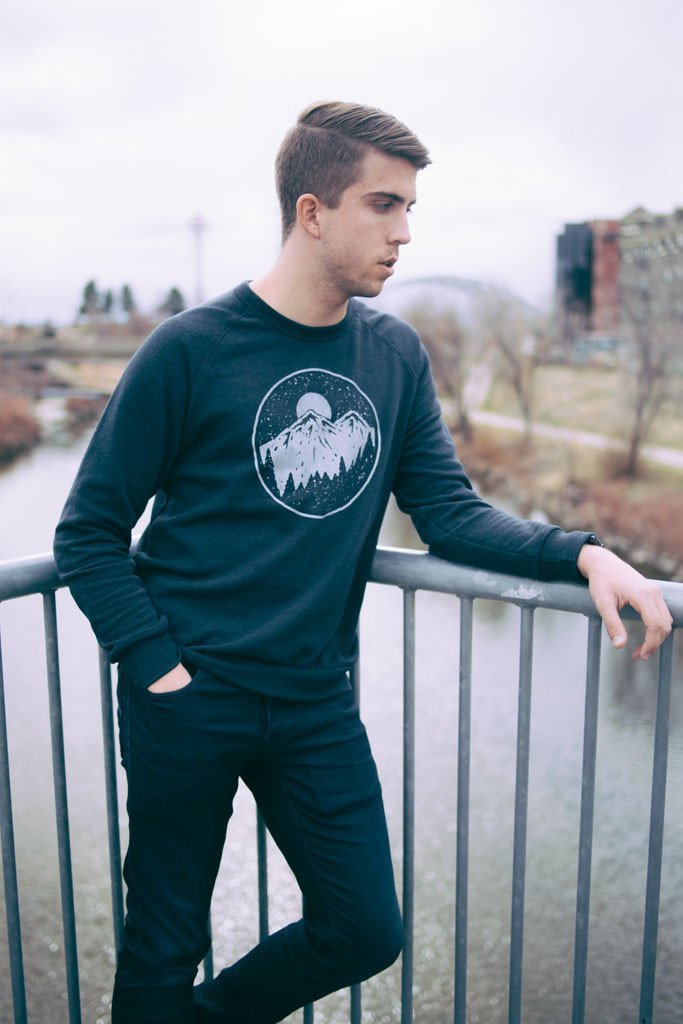 Man wearing black crew neck sweatshirt featuring moon over mountains graphic, leaning on railing overlooking a river.