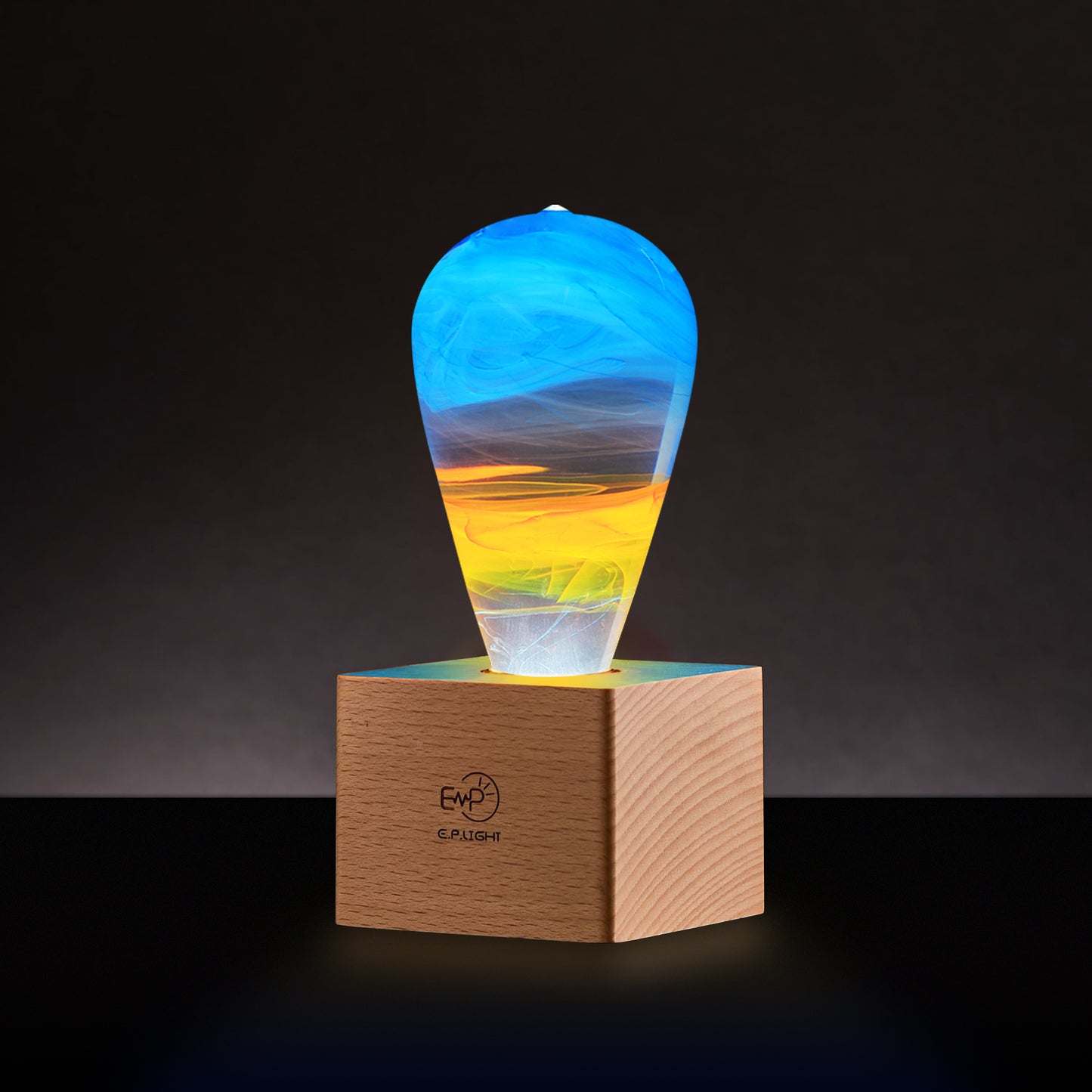 Clear lightbulb glowing from within, filled with yellow, orange, and blue swirls, set in cube shaped wood base in dim room.