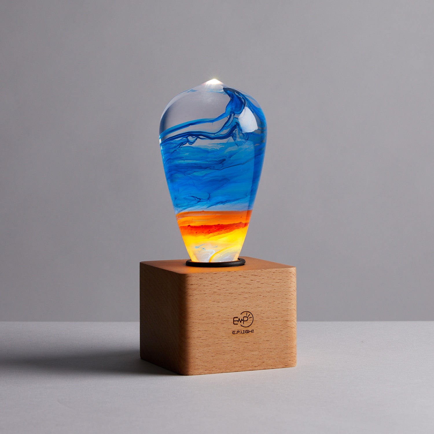 Clear lightbulb glowing from within, filled with yellow, orange, red, and blue swirls, set in cube shaped wood base.