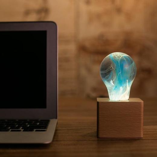 Clear lightbulb glowing with blue and yellow swirls, set in wooden base atop wood desk, beside silver laptop.