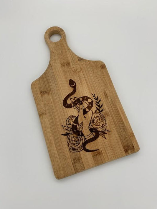 Bamboo paddle shaped cutting/serving board, burned in design of a hand holding a snake, surrounded by flowers.