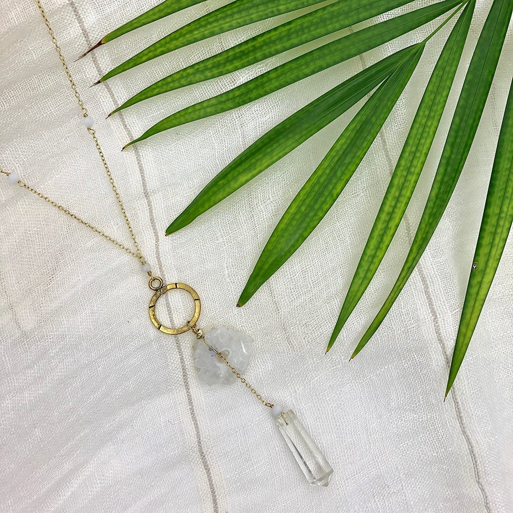 Y necklace with circular pendant, blow hangs solar quartz stone, and clear crystal, on brass chain, shown on cloth with leaf.